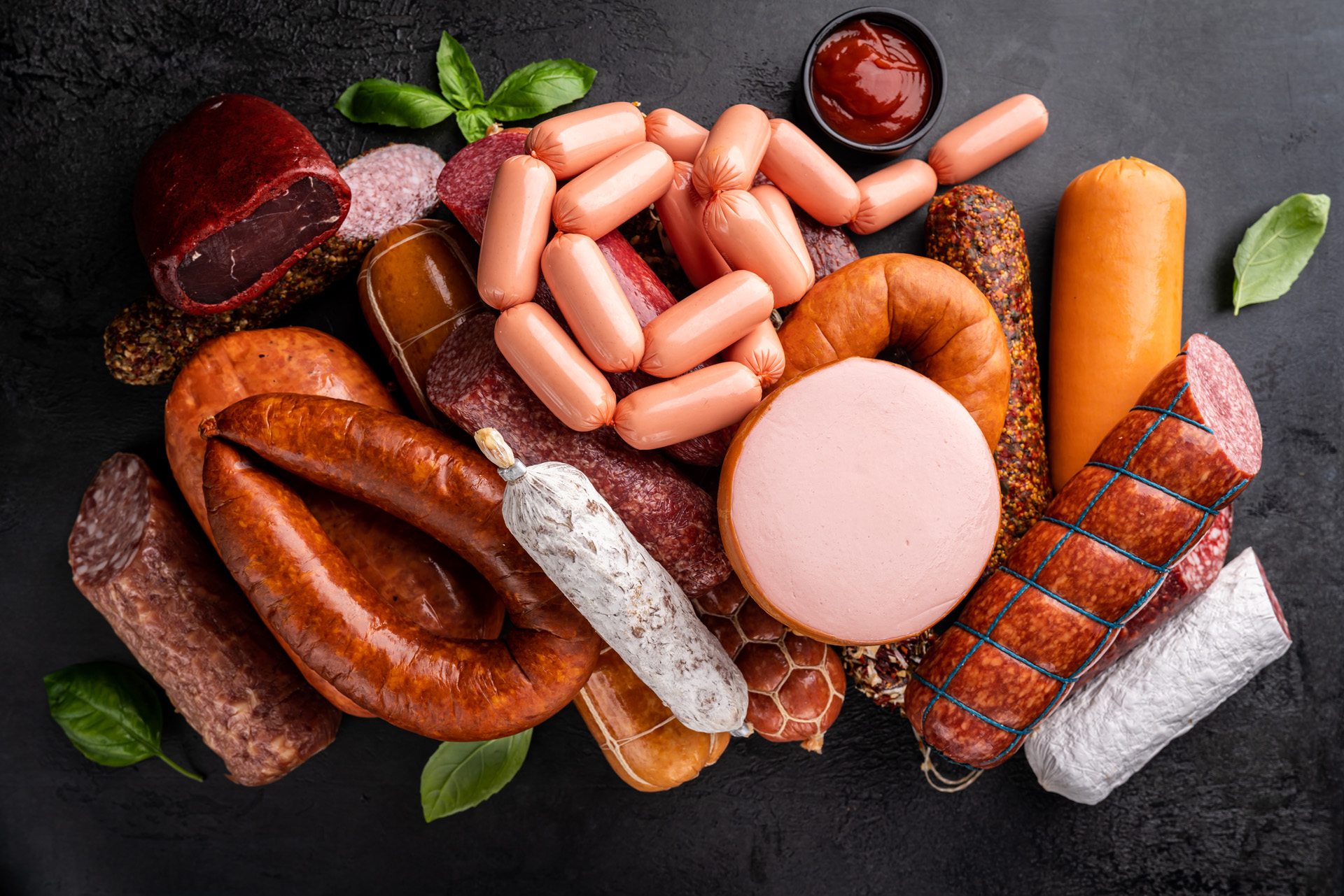 Set of different types of sausages, salami and smoked meat with basil and spices on a black stone background. Top view.