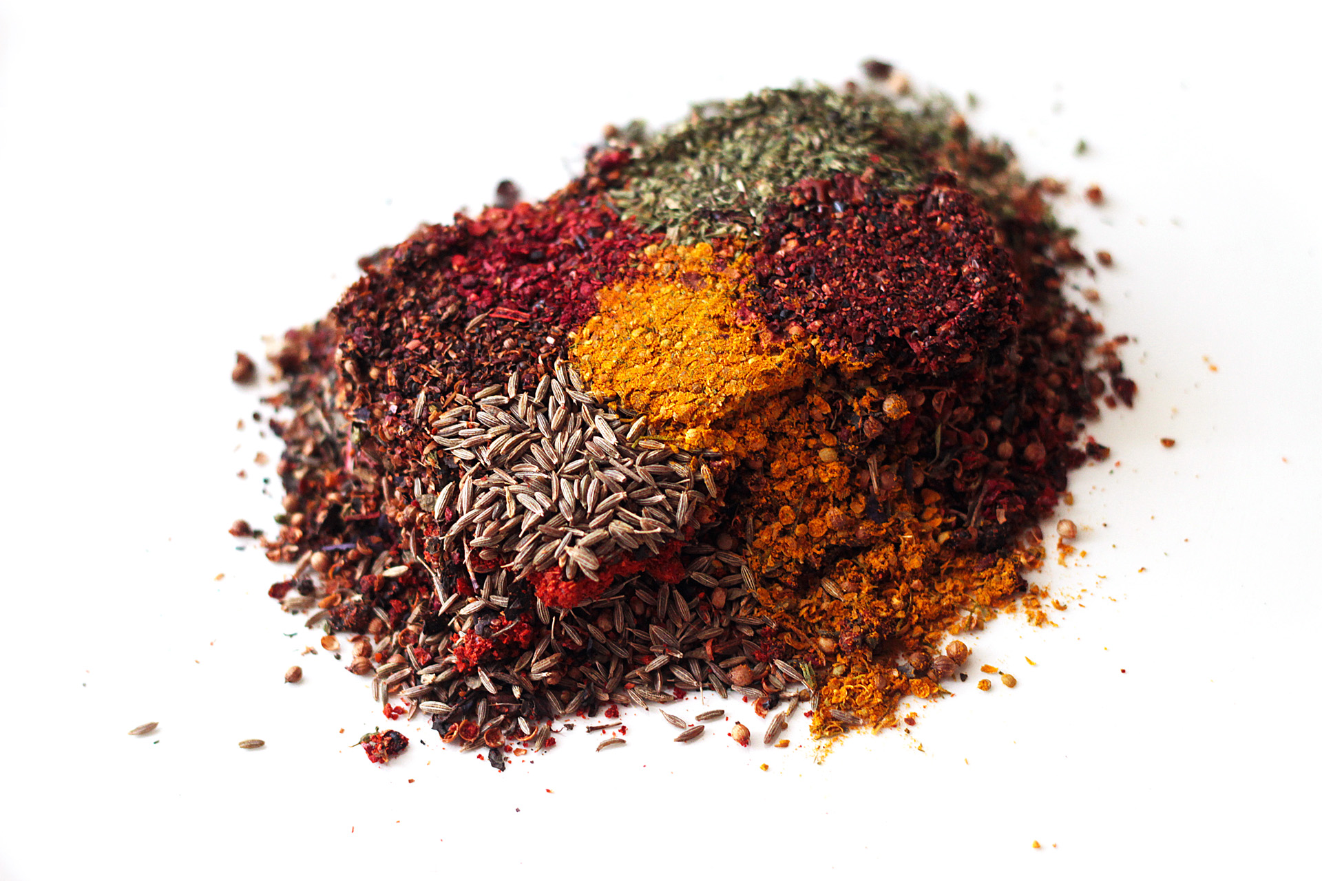 handful of separated spices of coriander, cumin, turmeric, dried tomato, sweet pepper, basil, jambil, black pepper on white background